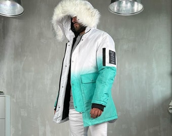 Pode incluir: Um parka gradiente branco e verde-azulado com capuz de pele sintética. O casaco tem um fecho de correr preto e um patch na manga. A pessoa está a usar calças de ganga brancas e ténis estampados.