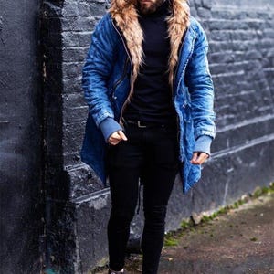 May include: A blue denim parka with a large tan faux fur hood lining, a black turtleneck sweater, black slim-fit trousers, and black and white low-top sneakers. The jacket features ribbed gray cuffs and a front zipper closure.