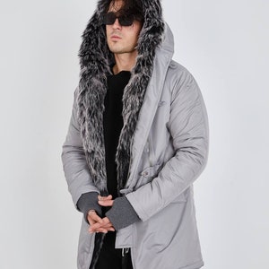 May include: A light gray parka featuring a large hood lined with thick, dark gray and white faux fur. The jacket has a smooth, matte finish, long sleeves with ribbed gray cuffs, and a front zipper closure. It is worn over a black sweater.