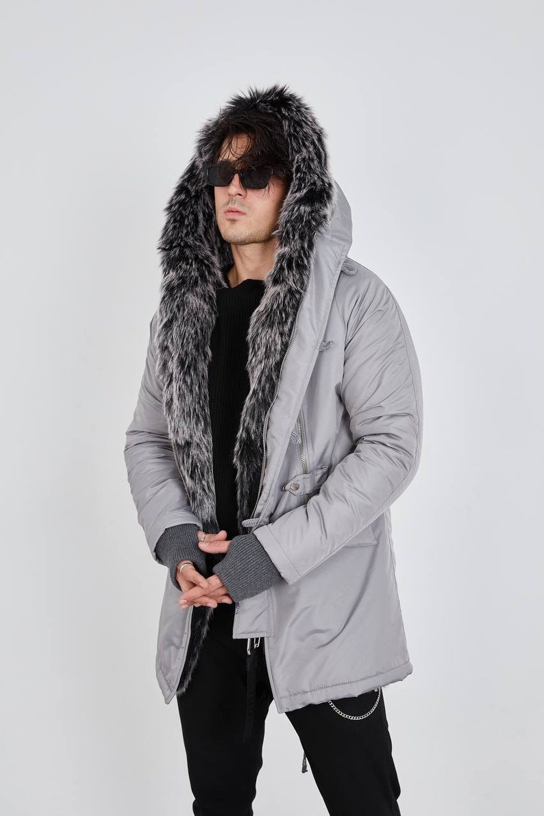 May include: A light gray parka featuring a large hood lined with thick, dark gray and white faux fur. The jacket has a smooth, matte finish, long sleeves with ribbed gray cuffs, and a front zipper closure. It is worn over a black sweater.
