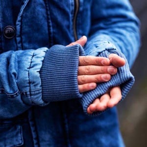 May include: A blue denim jacket featuring a dark wash, a prominent metal zipper closure, and a black button detail on the front. The sleeves are finished with wide, ribbed blue knit cuffs that extend past the wrist, providing a textured contrast.