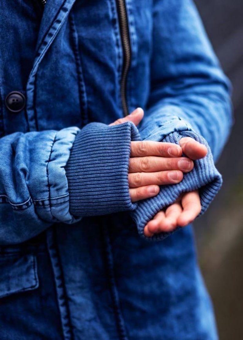 May include: A blue denim jacket featuring a dark wash, a prominent metal zipper closure, and a black button detail on the front. The sleeves are finished with wide, ribbed blue knit cuffs that extend past the wrist, providing a textured contrast.