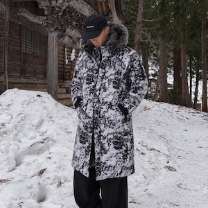 May include: A long, knee-length winter coat featuring a black and white abstract camouflage pattern, a hood with a thick grey faux fur trim, and black ribbed cuffs. The coat is worn with black trousers and a black baseball cap with white text.