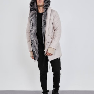 May include: A beige parka featuring a large hood lined with thick, dark grey and white faux fur. The coat has a front zipper closure, long sleeves with beige ribbed cuffs, and two front pockets. It is worn over a black sweater and black pants.