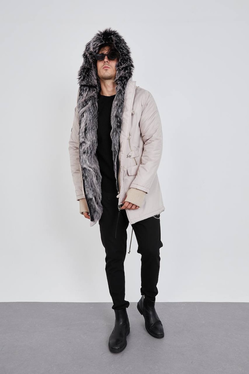 May include: A beige parka featuring a large hood lined with thick, dark grey and white faux fur. The coat has a front zipper closure, long sleeves with beige ribbed cuffs, and two front pockets. It is worn over a black sweater and black pants.