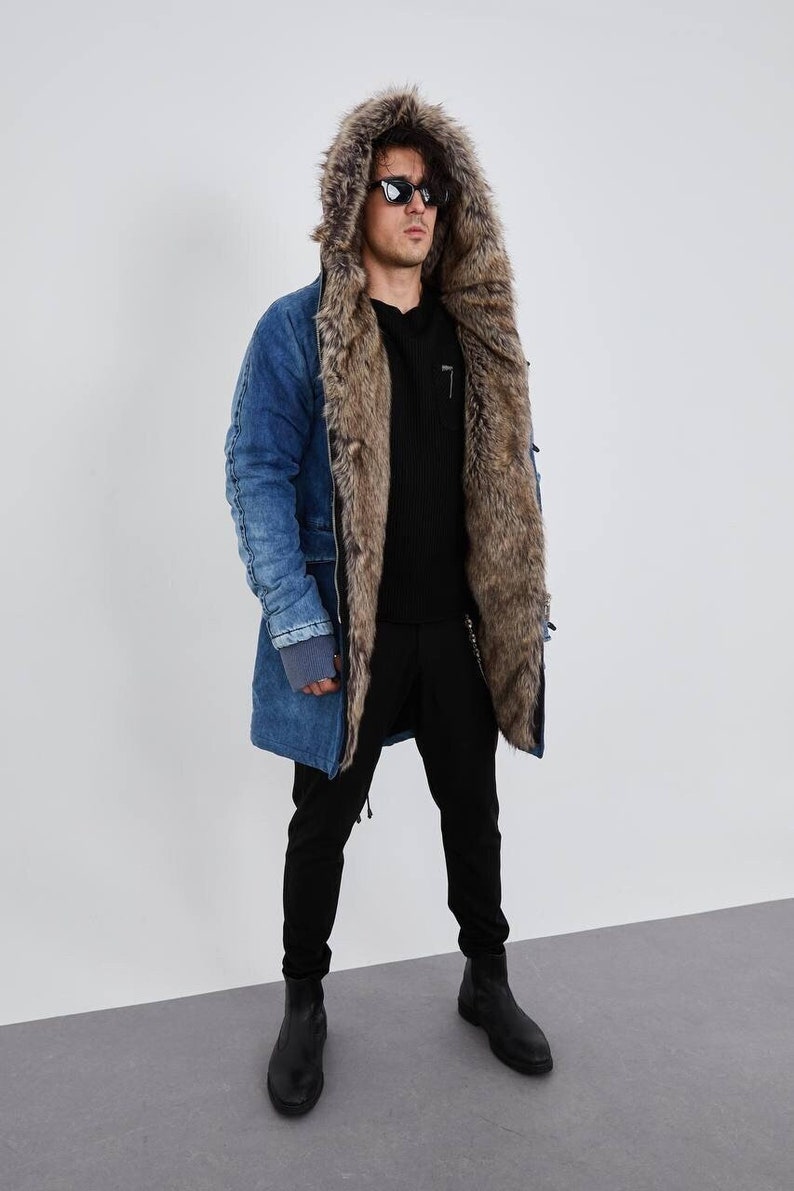May include: A blue denim parka featuring a large hood lined with thick brown faux fur, a front zipper closure, and ribbed gray cuffs. The jacket is worn over a black long-sleeved shirt and black trousers, paired with black leather boots.