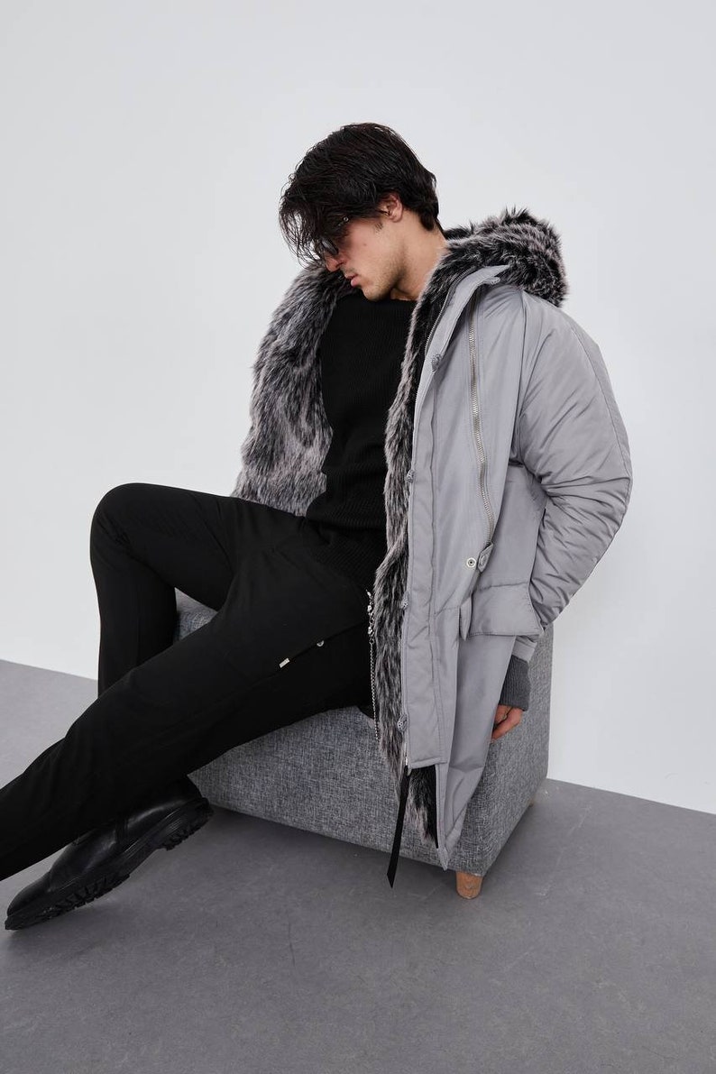 May include: A light gray parka featuring a hood lined with dark gray and white faux fur, a front zipper closure, and long sleeves with ribbed cuffs. The coat is worn over a black long-sleeved sweater and black trousers with black leather shoes.
