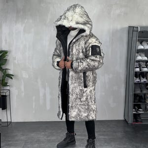 May include: A long, knee-length coat featuring a grey and white faux fur exterior with a large hood. The coat includes a black front zipper closure, black ribbed cuffs, and a small black utility pocket on the left sleeve with a circular emblem.