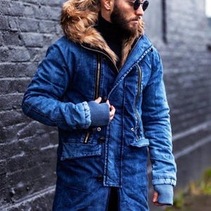May include: A blue denim parka featuring a hood with a thick brown faux fur trim, long sleeves with ribbed cuffs, vertical zippered chest pockets, and a button-down front closure. The jacket has a structured collar and a mid-thigh length design.
