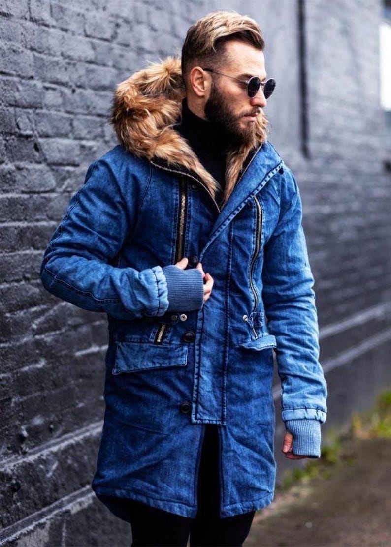 May include: A blue denim parka featuring a hood with a thick brown faux fur trim, long sleeves with ribbed cuffs, vertical zippered chest pockets, and a button-down front closure. The jacket has a structured collar and a mid-thigh length design.