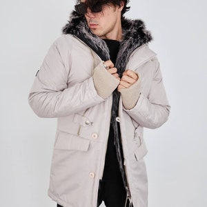 May include: A light beige parka jacket featuring a hood lined with dark gray and white faux fur, a front zipper closure, two flap pockets, and beige ribbed knit cuffs. The jacket is worn over a black sweater and black trousers.