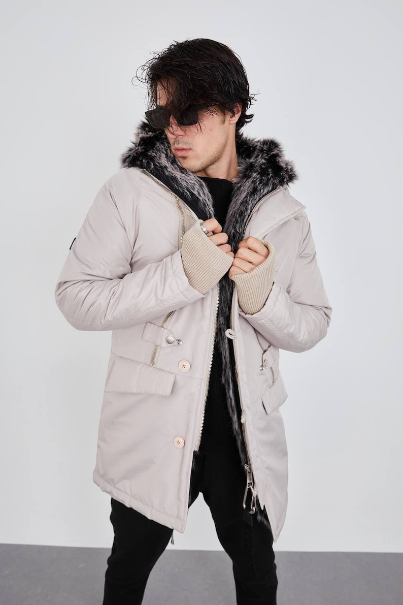 May include: A light beige parka jacket featuring a hood lined with dark gray and white faux fur, a front zipper closure, two flap pockets, and beige ribbed knit cuffs. The jacket is worn over a black sweater and black trousers.