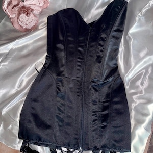 May include: A black satin corset with a sweetheart neckline and a front zipper closure. The corset has boning for structure and garter straps attached at the bottom. A decorative pink flower is in the background.