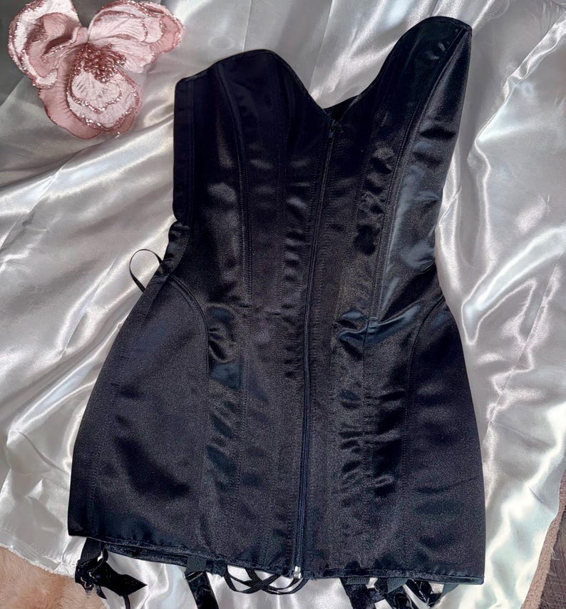 May include: A black satin corset with a sweetheart neckline and a front zipper closure. The corset has boning for structure and garter straps attached at the bottom. A decorative pink flower is in the background.