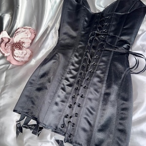 May include: A black satin corset with a lace-up front and garter straps. The corset is a classic design, featuring boning for structure and a smooth, shiny finish. The garment is displayed on a white, silky surface.