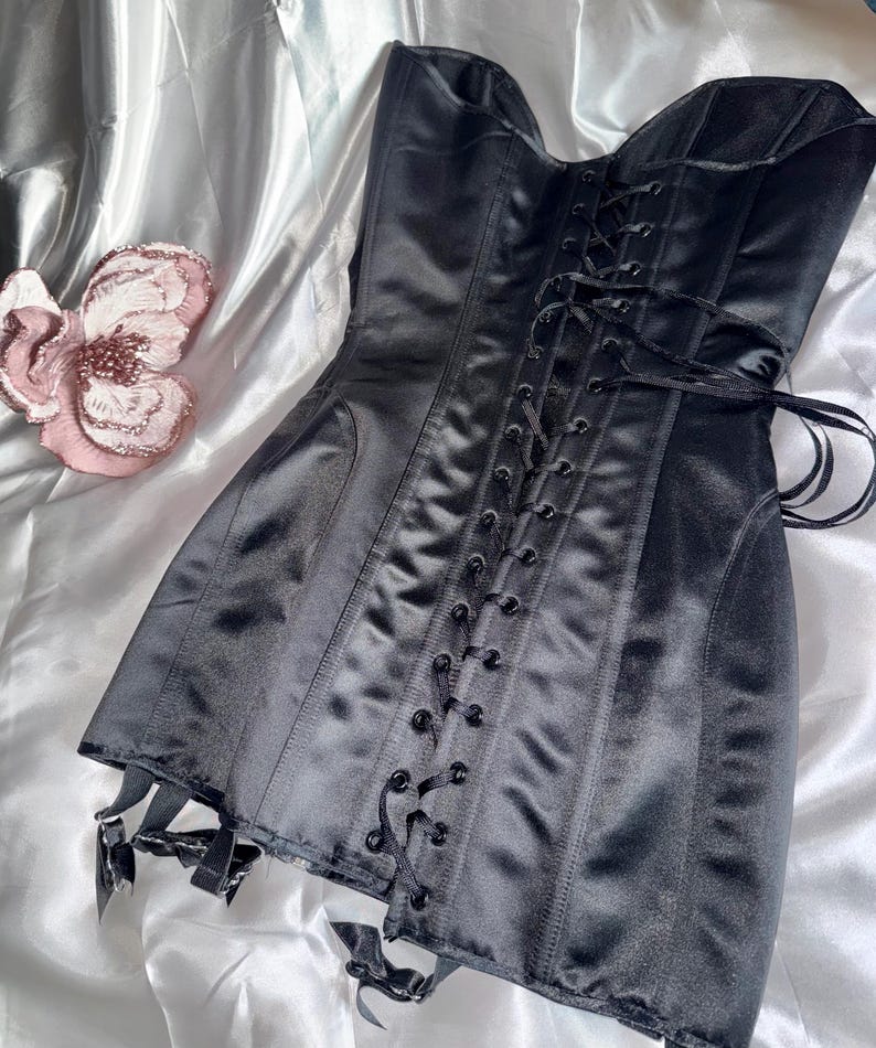 May include: A black satin corset with a lace-up front and garter straps. The corset is a classic design, featuring boning for structure and a smooth, shiny finish. The garment is displayed on a white, silky surface.