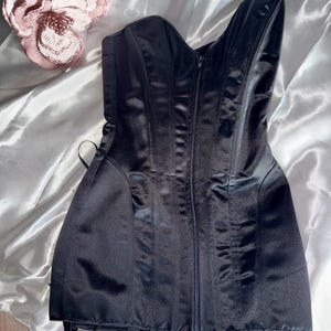 May include: A black satin corset with a zipper closure down the front. The corset has boning for structure and adjustable straps. The garment is displayed on a white satin surface, with a decorative pink flower in the upper left corner.