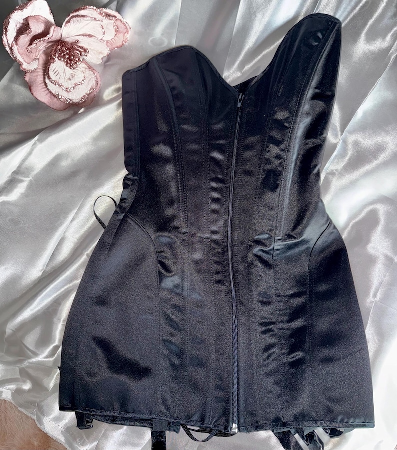 May include: A black satin corset with a zipper closure down the front. The corset has boning for structure and adjustable straps. The garment is displayed on a white satin surface, with a decorative pink flower in the upper left corner.