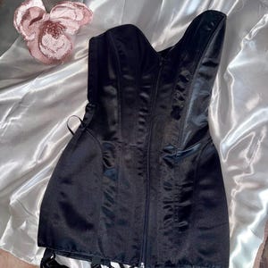 May include: A black satin corset with a zipper closure down the front. The corset has boning for structure and garter straps at the hem. A decorative pink flower is in the background. This garment is a lingerie item.