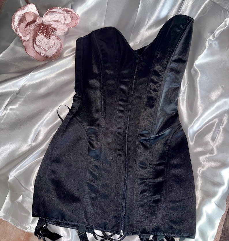 May include: A black satin corset with a zipper closure down the front. The corset has boning for structure and garter straps at the hem. A decorative pink flower is in the background. This garment is a lingerie item.