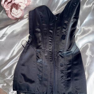 May include: A black satin corset with a zipper closure down the front. The corset has vertical seams and a sweetheart neckline. The garment is displayed on a white satin surface, with a pink flower in the upper left corner.