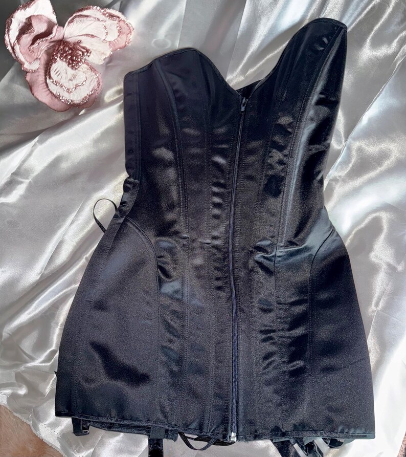 May include: A black satin corset with a zipper closure down the front. The corset has vertical seams and a sweetheart neckline. The garment is displayed on a white satin surface, with a pink flower in the upper left corner.