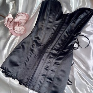 May include: A black satin corset with a zipper closure and boning details. The corset has a sweetheart neckline and is laced in the back. A pink flower is in the background.