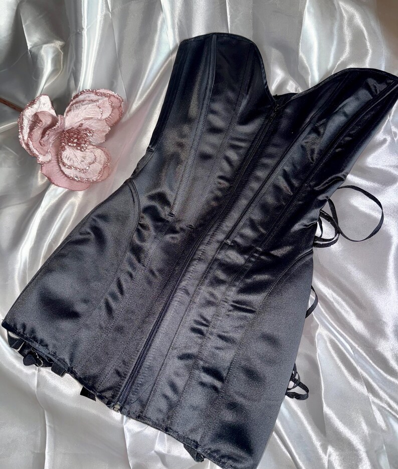 May include: A black satin corset with a zipper closure and boning details. The corset has a sweetheart neckline and is laced in the back. A pink flower is in the background.