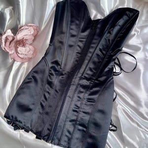 May include: A black satin corset with a zipper closure and boning details. The corset has a sweetheart neckline and a lace-up back. A pink flower decoration is visible on the left side of the image.