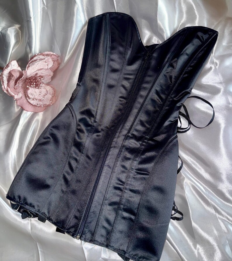 May include: A black satin corset with a zipper closure and boning details. The corset has a sweetheart neckline and a lace-up back. A pink flower decoration is visible on the left side of the image.
