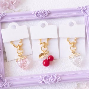 May include: Three decorative keychains with gold-colored hardware and charms. One keychain features a pink strawberry charm, another has a red cherry charm, and the third has a clear strawberry charm. The keychains are displayed in a lavender picture frame.