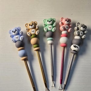 May include: Five metal dotting tools with textured handles, each topped with a silicone bear figure. The bears are light blue, tan, mint green, pink, and gray. The tools feature colorful silicone beads in various shapes and sizes below the bear figures.
