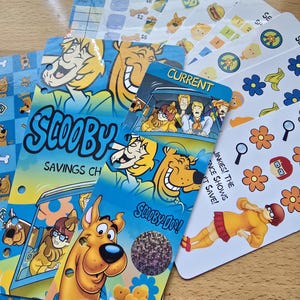 May include: A collection of Scooby-Doo themed savings challenge trackers and sticker sheets. The items feature colorful illustrations of Scooby-Doo, Shaggy, and Velma, with text including "SCOOBY SAVINGS CHALLENGE," "CURRENT," and "JINKIES! THE EVIDENCE SHOWS IT SAVE!"