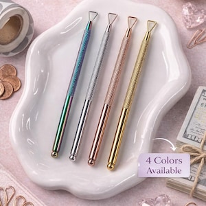 Puede incluir: Cuatro herramientas para raspar en iridiscente, plateado, oro rosa y dorado, presentadas en un plato blanco ondulado. Las herramientas tienen mangos texturizados y puntas triangulares. La imagen incluye el texto "4 Colores Disponibles" y "Perfecto para desafíos de ahorro."