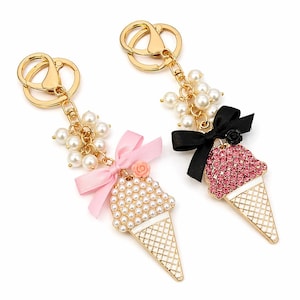 Könnte beinhalten: Zwei goldfarbene Eiswaffel-Schlüsselanhänger. Einer hat eine rosa Schleife und Perlen, der andere eine schwarze Schleife und rosa Edelsteinakzente. Beide haben goldene Schlüsselringe und Perlen.
