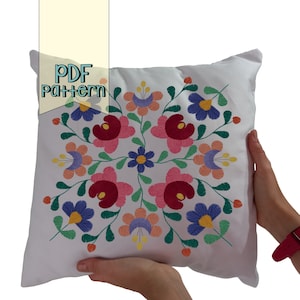 Puede incluir: Almohada blanca con un diseño floral bordado de colores. El diseño presenta flores rojas, rosas, azules, verdes y amarillas.