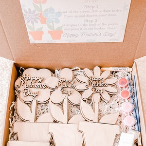 Puede incluir: Un kit de bricolaje para el Día de la Madre, con recortes de flores de madera y el texto "Happy Mother's Day". El kit incluye pintura, pinceles e instrucciones dentro de una caja de cartón. El kit está diseñado para ser pintado y ensamblado.