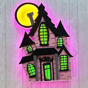Puede incluir: Decoración de casa embrujada rosa iluminada con ribetes negros y un fondo de luna llena. Las ventanas y la puerta brillan en verde, y la luna brilla en amarillo. La casa tiene múltiples torretas y una chimenea.