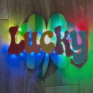 May include: A vibrant "LUCKY" sign in a retro font, set on a striped shamrock shape. The sign is illuminated with blue, green, and red lights, creating a colourful display. The background is a wooden surface.