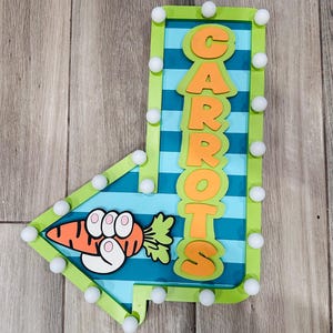 May include: A decorative arrow-shaped sign with the word "CARROTS" in orange letters. The sign features a cartoon carrot with a hand holding it. The sign is outlined in green with white ball-shaped lights.
