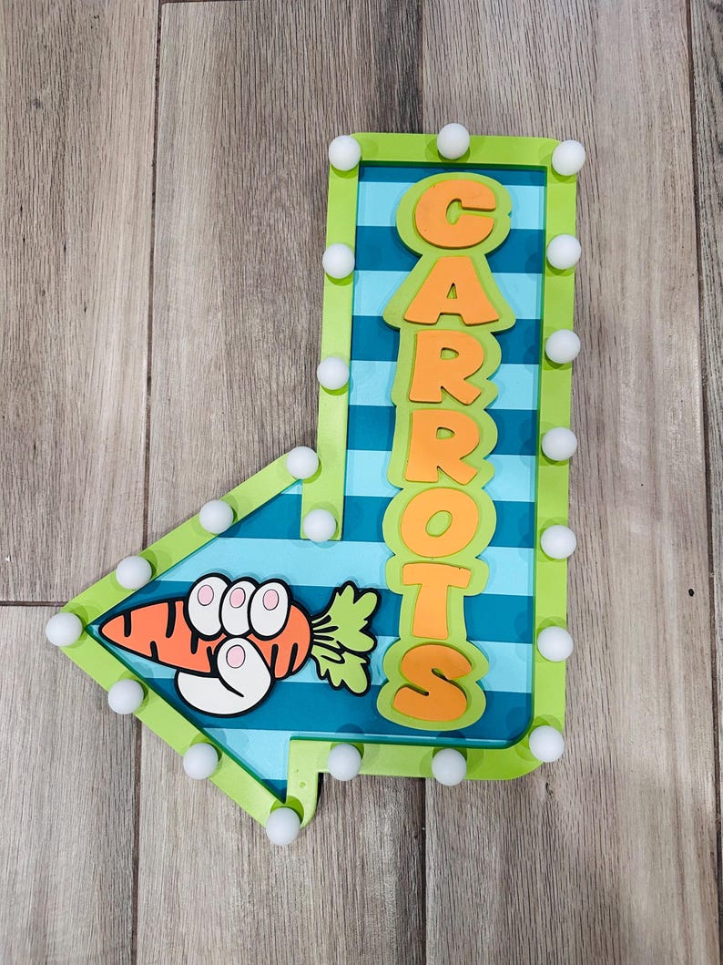 May include: A decorative arrow-shaped sign with the word "CARROTS" in orange letters. The sign features a cartoon carrot with a hand holding it. The sign is outlined in green with white ball-shaped lights.