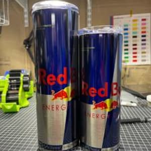 May include: Two stainless steel tumblers with a Red Bull design. The tumblers are blue with the Red Bull logo in red and yellow. The tumblers have a silver band at the bottom and a clear lid. One tumbler is taller than the other.