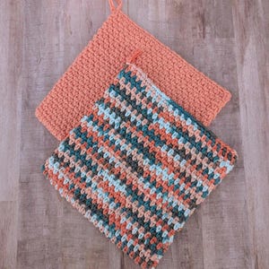Puede incluir: Dos agarraderas de olla de ganchillo, una en color melocotón sólido y la otra en un patrón de rayas multicolores de melocotón, azul verdoso y marrón.