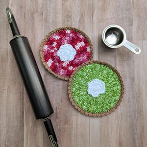 May include: A kitchen scene featuring a black rolling pin, two crocheted coasters, and a measuring cup. The coasters are round, one red and pink, the other green and white. The rolling pin is dark grey.