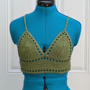 May include: A green crocheted crop top with a triangular neckline and thin straps. The top is made with a simple stitch pattern and has a delicate, feminine look.
