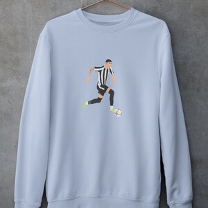 Puede incluir: Una sudadera de color azul claro con un gr&aacute;fico de un jugador de f&uacute;tbol con una camiseta a rayas blancas y negras regateando un bal&oacute;n de f&uacute;tbol.