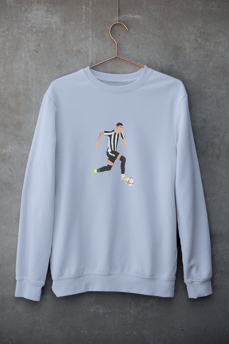 Puede incluir: Una sudadera de color azul claro con un gr&aacute;fico de un jugador de f&uacute;tbol con una camiseta a rayas blancas y negras regateando un bal&oacute;n de f&uacute;tbol.