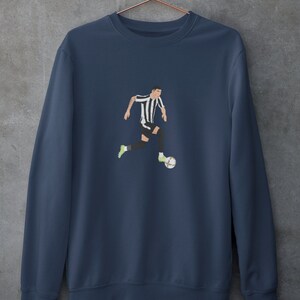 Puede incluir: Una sudadera azul marino con un gr&aacute;fico de un jugador de f&uacute;tbol con una camiseta negra y blanca regateando un bal&oacute;n de f&uacute;tbol.