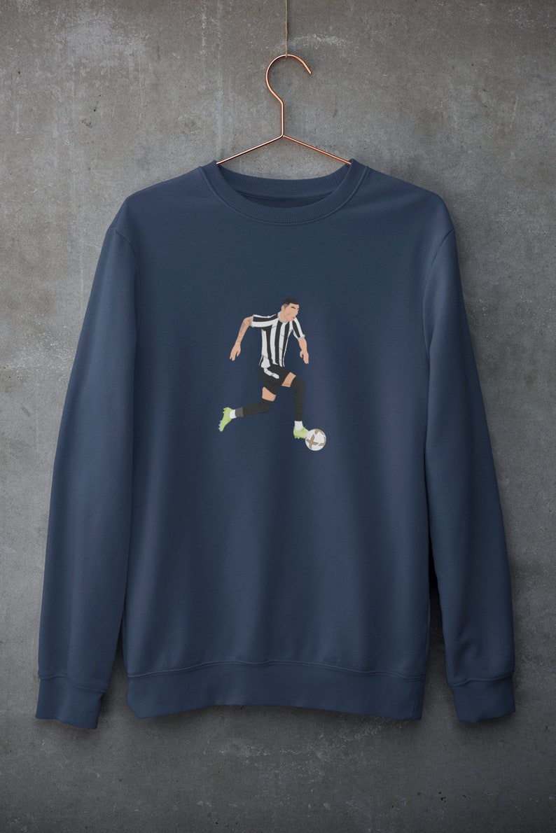 Puede incluir: Una sudadera azul marino con un gr&aacute;fico de un jugador de f&uacute;tbol con una camiseta negra y blanca regateando un bal&oacute;n de f&uacute;tbol.