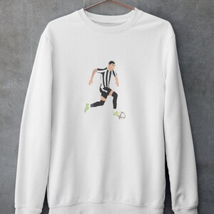 Puede incluir: Sudadera blanca con una ilustraci&oacute;n en blanco y negro de un jugador de f&uacute;tbol driblando el bal&oacute;n.